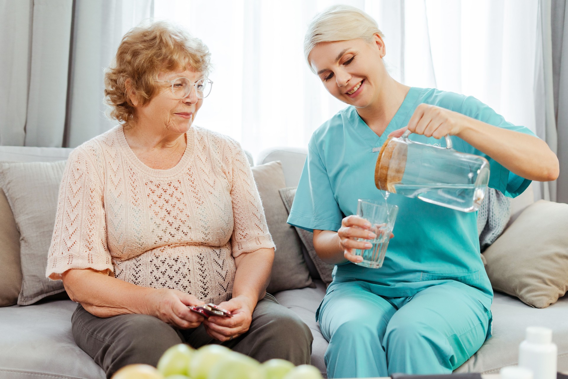 Smiling nurse pouring glass of water for elderly woman on sofa